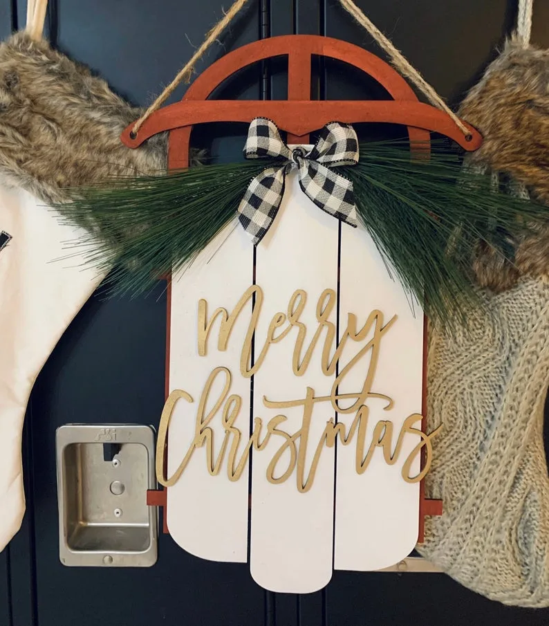 A wooden 'Merry Christmas' sled with a buffalo check bow and pine sprigs, hanging on a navy front door next to a stocking